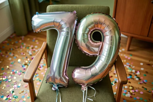 Twee grote metallic ballonnen in de vorm van een 7 en een 9 zijn vastgemaakt aan een groen beklede stoel met houten armleuningen, boven een houten vloer met kleurrijke confetti.