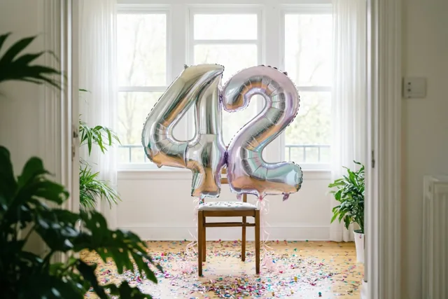 Grote zilver- en pastelkleurige cijferballonnen vormen 42 aan een houten stoel in een lichte kamer met confetti, witte gordijnen en groene planten.