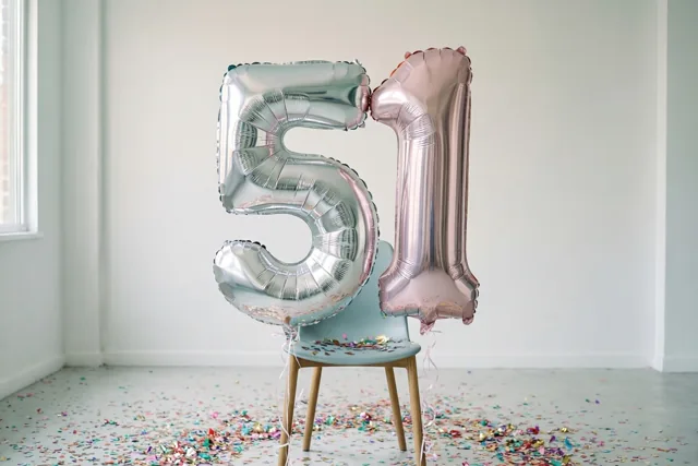 Zilveren en roségouden folieballonnen 5 en 1 zijn vastgemaakt aan een lichtblauwe stoel in een lichte, sober ingerichte kamer met kleurrijke confetti op de vloer.