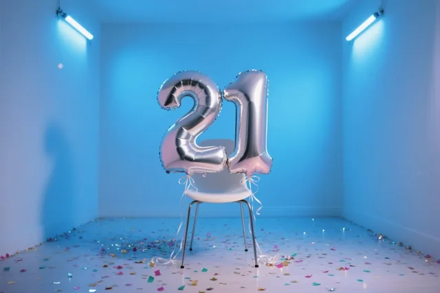 Zilveren folieballonnen vormen 21 aan een witte stoel in een blauwe kamer, met kleurrijke confetti op de vloer en felblauw licht van het plafond.