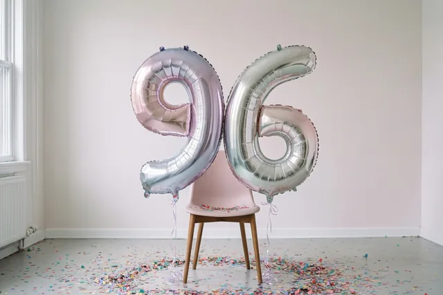 Grote folieballonnen in de vorm van een 9 en een 6 zijn vastgemaakt aan een roze stoel in een lichte, sobere kamer met confetti op de vloer en daglicht uit een raam.