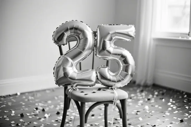 Zilveren cijferballonnen vormen 25 aan een stoel in een lichte kamer met confetti op de vloer, weergegeven in één kleurtoon.