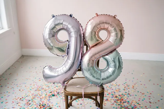 Twee grote folieballonnen 9 en 8 zijn aan een houten stoel vastgemaakt in een lichte kamer met zachtroze muren en kleurrijke confetti op de vloer.