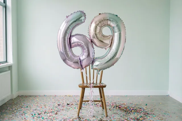 Twee grote zilveren en pastelkleurige cijferballonnen vormen 69 aan een houten stoel in een lichte kamer met mintgroene muren en kleurrijke confetti op de vloer.
