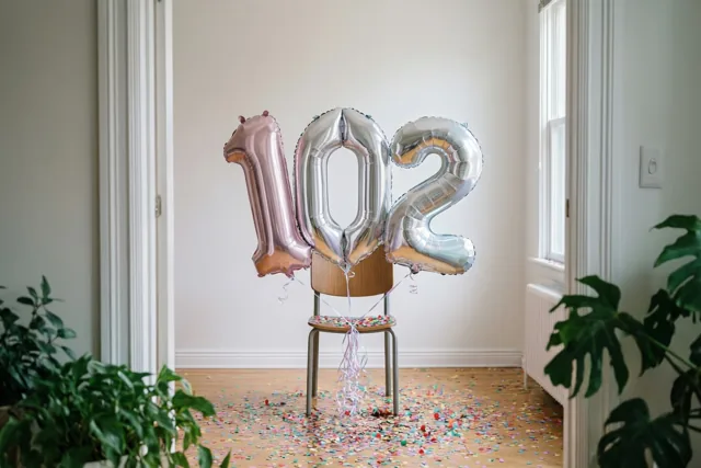 Folieballonnen in de vorm van 102 hangen aan een houten stoel in een lichte kamer met confetti op de houten vloer en groene planten aan beide kanten.
