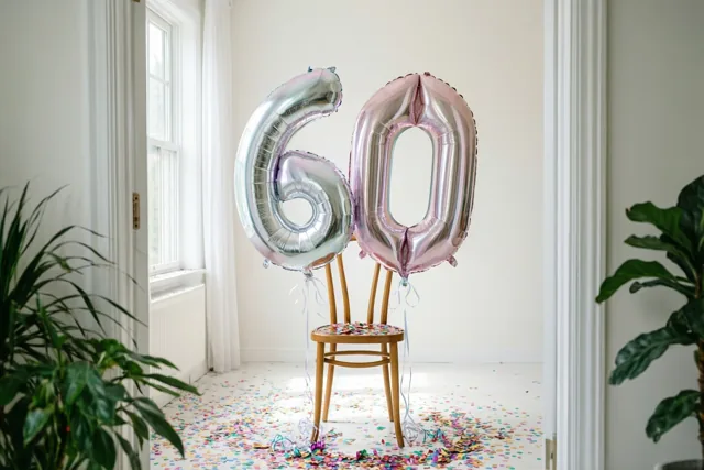 Folieballonnen in de vorm van 60 zijn aan een houten stoel vastgemaakt in een lichte kamer, met confetti op de vloer en planten naast de deuropening.