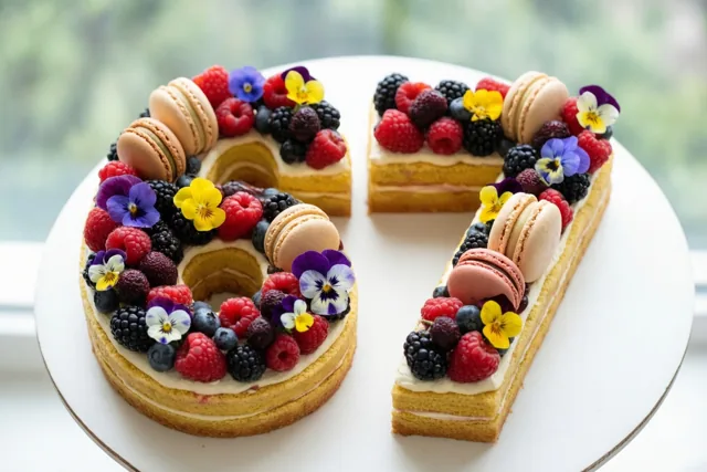 Een taart in de vorm van 67 met frambozen, bramen, macarons en eetbare bloemen staat op een wit bord tegen een onscherpe groene achtergrond.