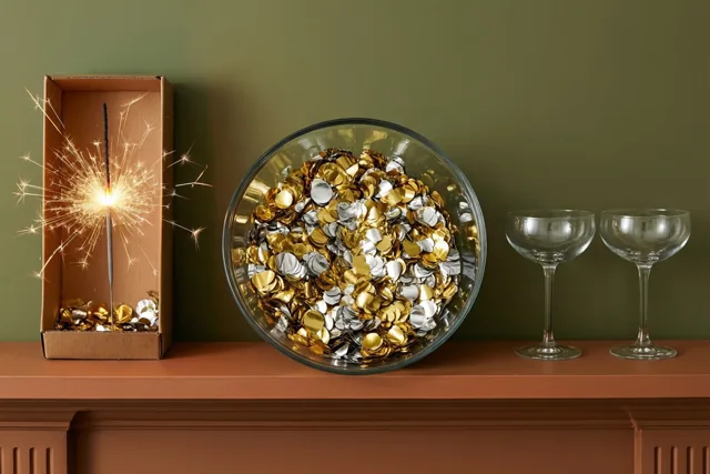 Een glazen schaal met gouden en zilveren confetti staat op een schoorsteenmantel naast een brandend sterretje in een kartonnen doos en twee lege champagnecoupes voor een groene muur.