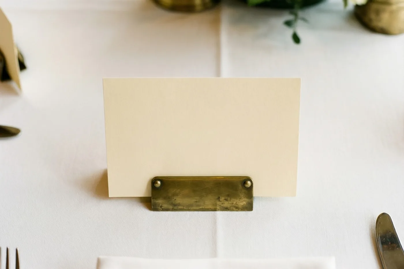 Een blanco crèmekleurig tafelkaartje staat rechtop tegen een messing plaatje op een wit tafelkleed, met deels zichtbaar bestek en bloemversiering op de achtergrond.