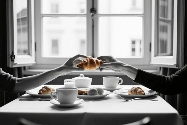 Twee handen reiken over een ontbijttafel om een croissant te delen, met een open raam op de achtergrond in zwart-wit.