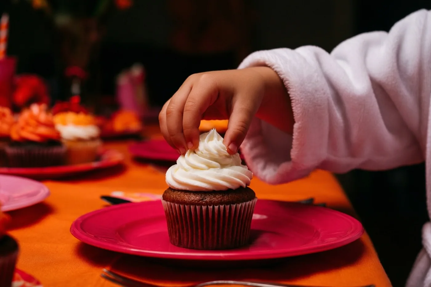 Een kinderhand uit een witte gewaadsmouw reikt naar een cupcake met wit glazuur op een roze bord, op een oranje tafelkleed met andere cupcakes op de achtergrond.