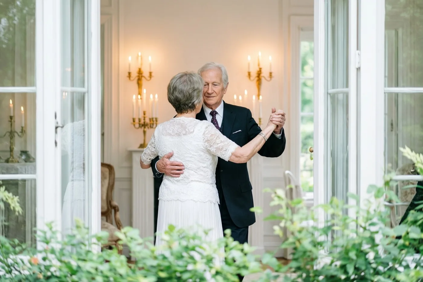 Een ouder koppel danst langzaam met de rug naar de kijker in een elegante kamer met brandende wandkandelaars, omlijst door witte openslaande deuren en groen op de voorgrond.
