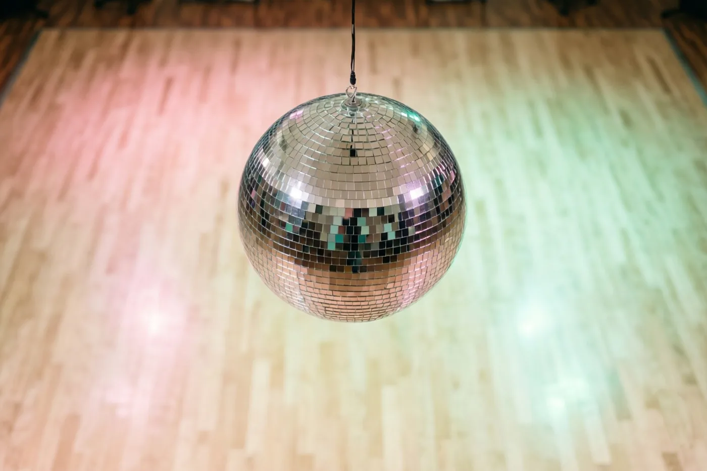 Een discobal hangt boven een lege houten dansvloer die door roze en groene lampen wordt verlicht.