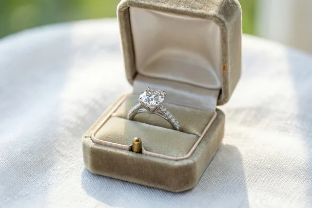 Een diamanten verlovingsring ligt in een open beige fluwelen doosje op een witte linnen doek in zacht daglicht.