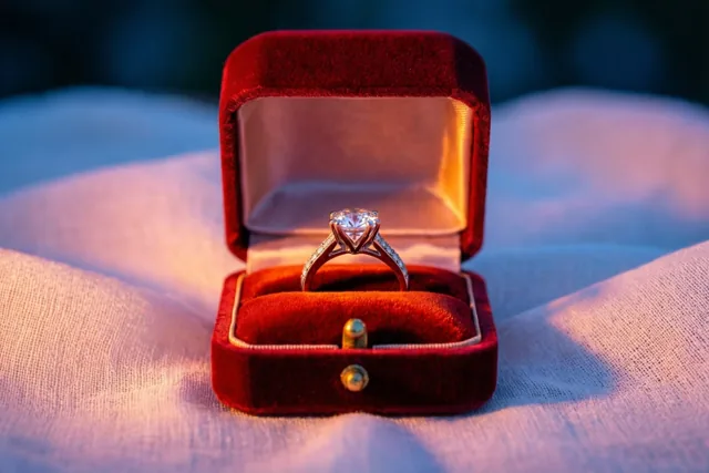 Een open rood fluwelen ringdoosje met een diamanten verlovingsring staat op een witte linnen doek in zacht, warm licht.