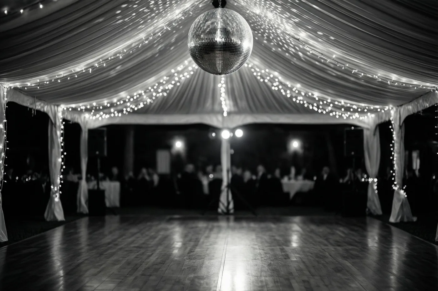 Een zwart-witfoto toont een lege houten dansvloer in een gedrapeerde feesttent met lichtslingers en een grote discobal die licht op de vloer weerkaatst.