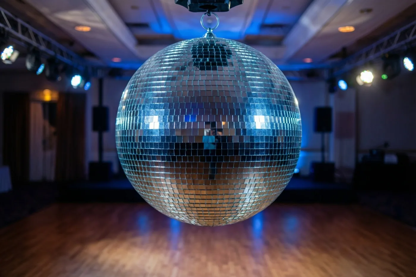 Een grote discobal hangt boven een glanzende houten dansvloer in een donkere feestzaal met blauw en warm licht.