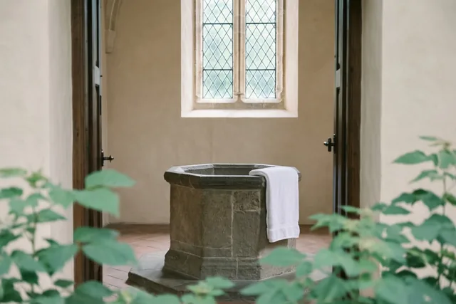 Een achthoekig stenen doopvont met een witte handdoek over de rand staat in een kerkruimte met licht door een glas-in-loodraam en groene bladeren op de voorgrond.