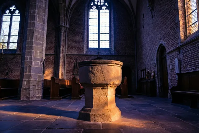 Een oude stenen doopvont staat in een schemerige Vlaamse dorpskerk met houten banken en bakstenen muren, verlicht door zonlicht uit een hoog raam.