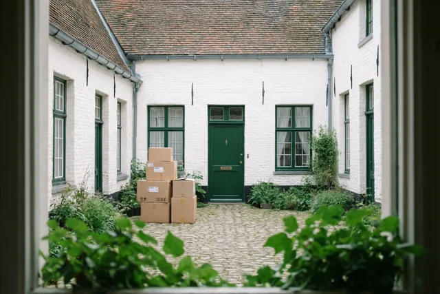 Door een raam is een rustig, gekasseid binnenhof in begijnhofstijl te zien, met witgekalkte gebouwen, groen en een stapel kartonnen verhuisdozen bij een groene deur.