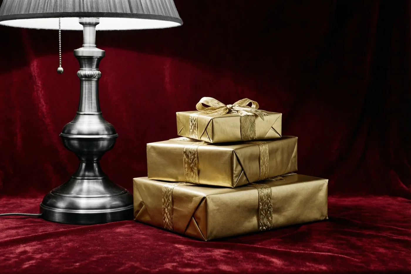 Drie goudkleurig ingepakte cadeaus staan op diep rood fluweel naast een brandende messing lamp.