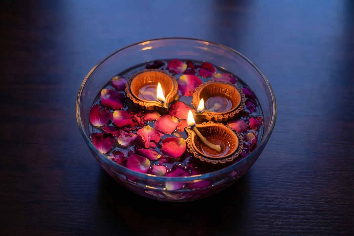 Een glazen schaal met water, drijvende rozenblaadjes en drie brandende olielampjes staat op een donkerhouten tafel voor een koele blauwe achtergrond.