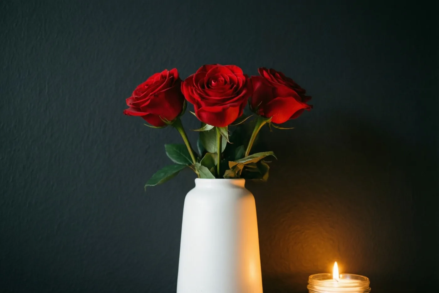 Drie rode rozen staan in een sobere witte keramieken vaas tegen een antracietkleurige muur, zacht verlicht door een kaars.