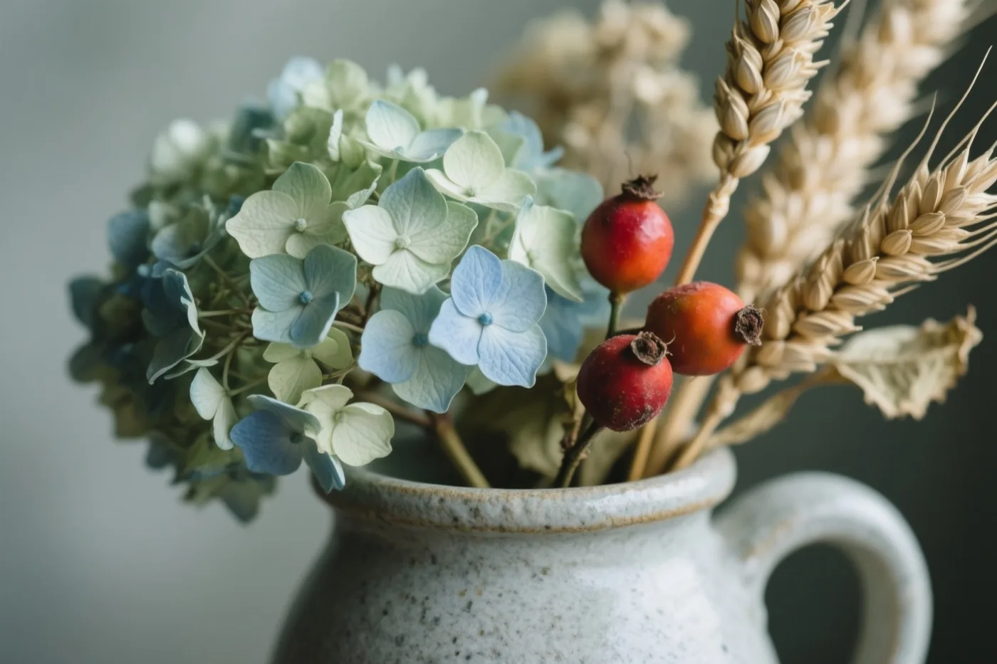 Gedroogde hortensia's, rode rozenbottels en tarwehalmen staan in een aardewerken kan tegen een rustige, neutrale achtergrond.