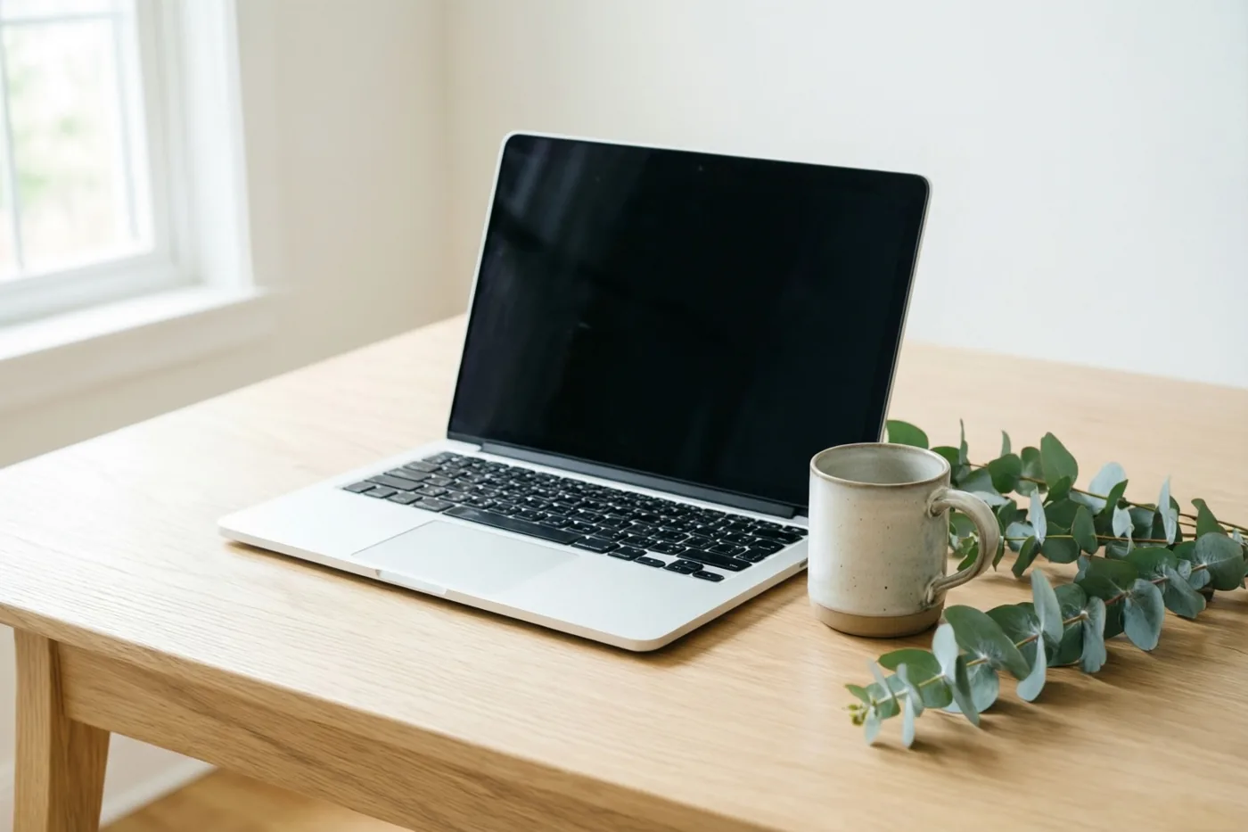 Een zilverkleurige laptop met zwart scherm staat op een licht eiken bureau naast een keramieken mok en een takje eucalyptus, in zacht licht van een raam.