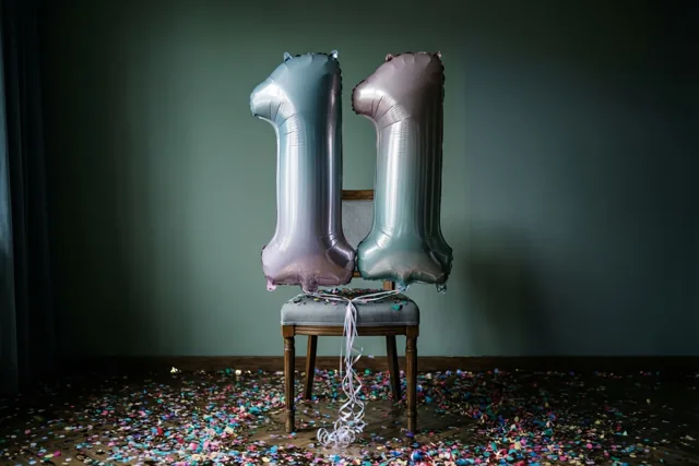 Twee grote folieballonnen vormen 11 aan een stoel voor een donkergroene muur, met confetti op de vloer en gedempte zilver- en pasteltinten.