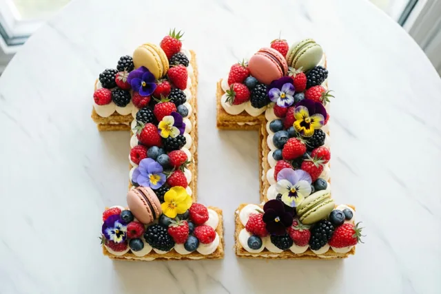Twee cijfertaarten vormen 11 op een wit marmeren oppervlak en zijn versierd met bessen, macarons en eetbare bloemen.