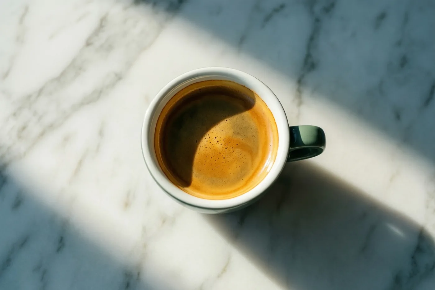 Een wit keramieken kopje espresso staat op een wit marmeren werkblad in vroeg zonlicht dat lange, zachte schaduwen werpt.