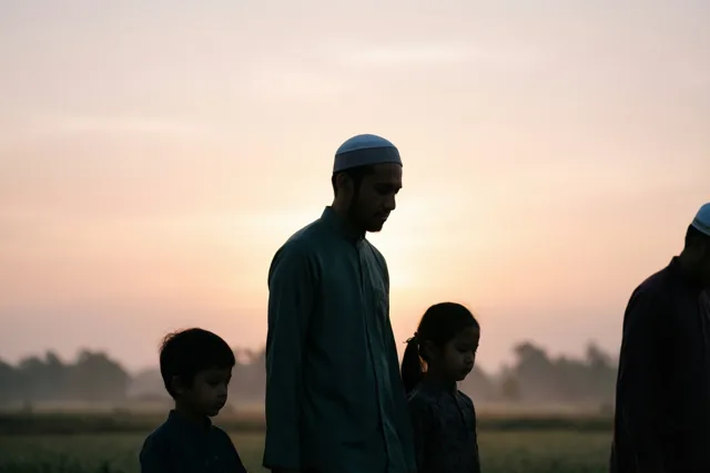 De silhouetten van een familie met volwassenen en kinderen lopen bij zonsopgang door een veld onder een nevelige roze en oranje lucht.