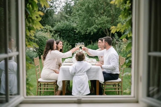 Door een raam en deels achter groen is een familie te zien die wijnglazen heft aan een witgedekte tuintafel, met een kind in een wit gewaad ertussen.