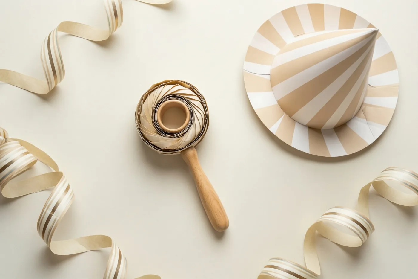 Een papieren feesthoedje en een ratel liggen tussen serpentines op een effen ondergrond, van bovenaf gefotografeerd in beige, wit en lichte houttinten.