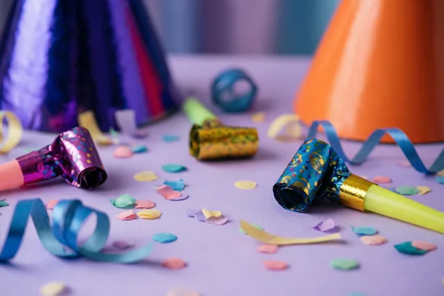Van bovenaf zie je feesthoedjes, roltongen, confetti en gekrulde linten verspreid over een pastelpaarse ondergrond.