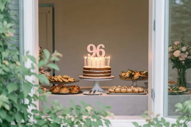 Door een open deuropening is een taart op een buffettafel te zien, met een papieren 96, brandende kaarsen en groen op de voorgrond.