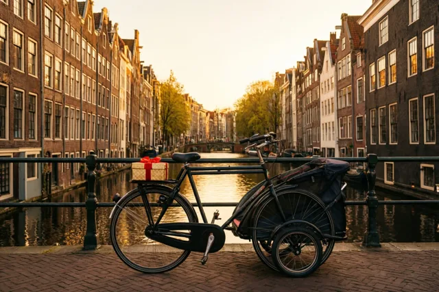 Een zwarte fiets met een kinderkar en een klein cadeaudoosje op het stuur staat op een bakstenen pad langs een kanaal met gevelhuizen, terwijl goudkleurig licht op het water weerspiegelt.