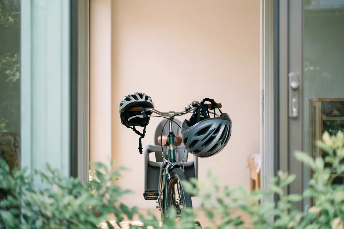 Een fiets met een kinderzitje en een grote en een kleine helm aan het stuur staat binnen bij beige muren en glazen deuren, gezien door groen op de voorgrond.