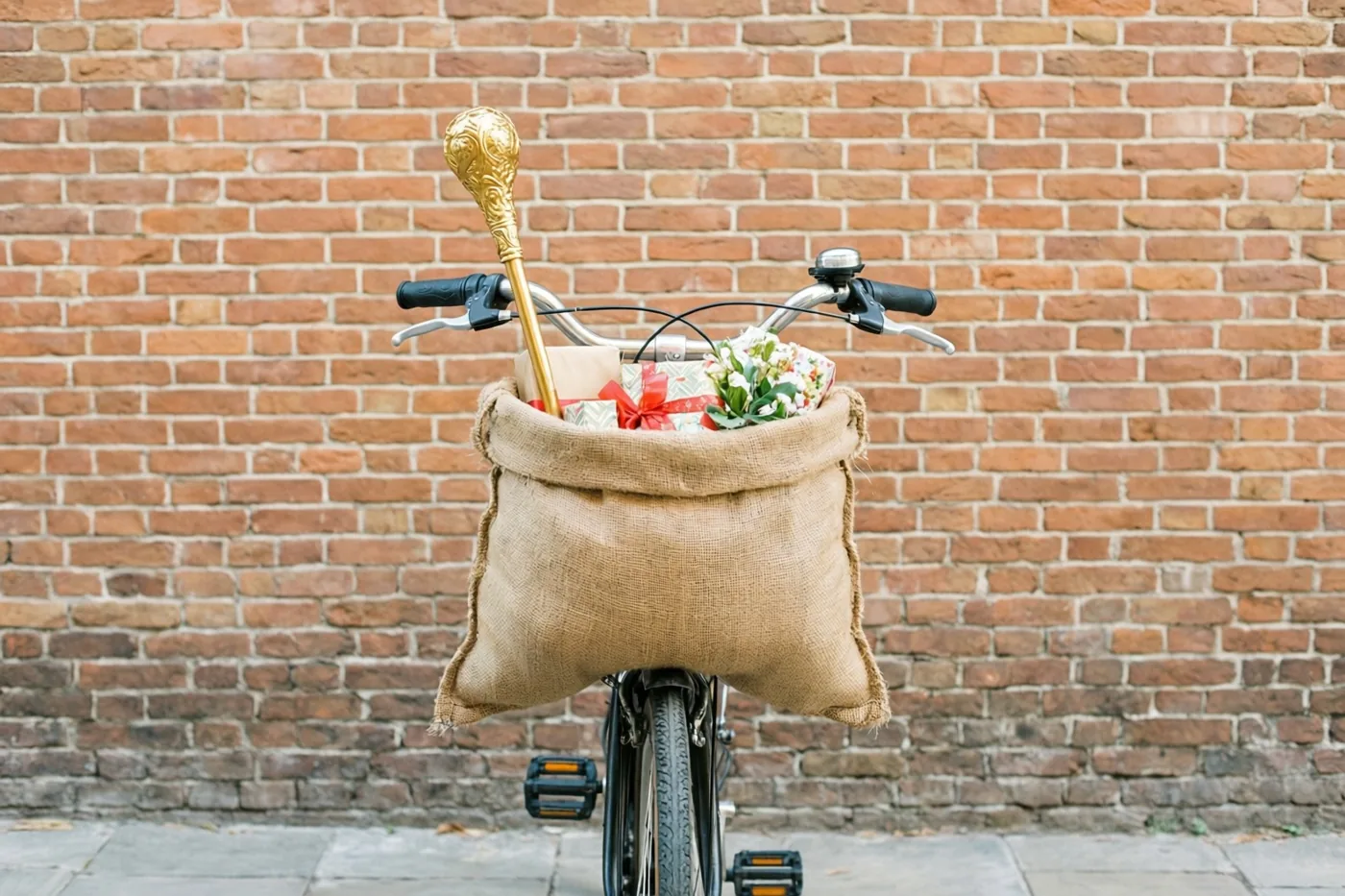 Een fiets met een jute zak vol ingepakte cadeaus en een gouden staf staat voor een rode bakstenen muur in warme tinten.