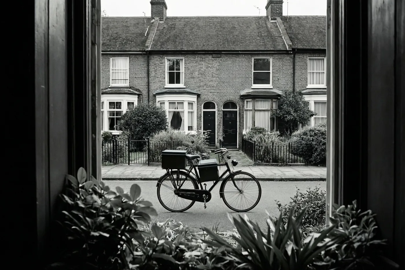 Een zwart-witfoto toont door een raam met planten een fiets met een doos op de bagagedrager voor een bakstenen rijhuis.