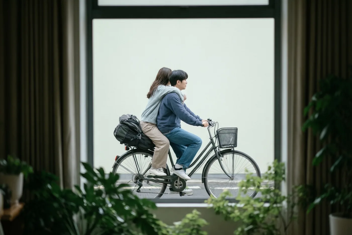 Twee mensen rijden samen op een fiets met één persoon op de bagagedrager, van achteren gezien door een raam met gordijnen en kamerplanten op de voorgrond.