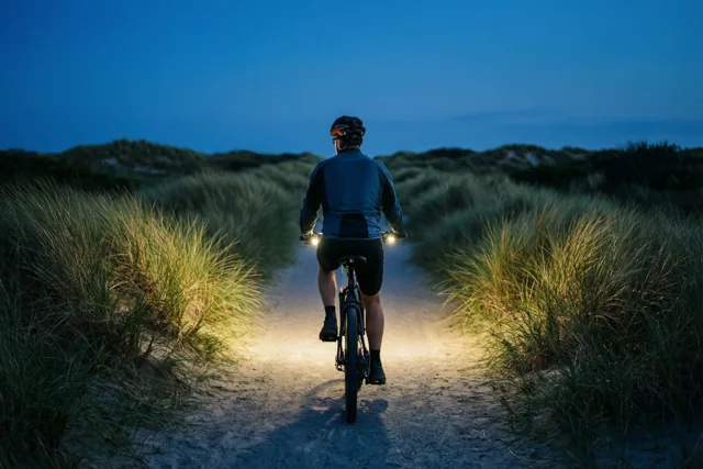 Een fietser rijdt van de kijker weg over een zandpad door de duinen, waar de fietslichten het pad en gras warm verlichten onder een diepblauwe avondhemel.