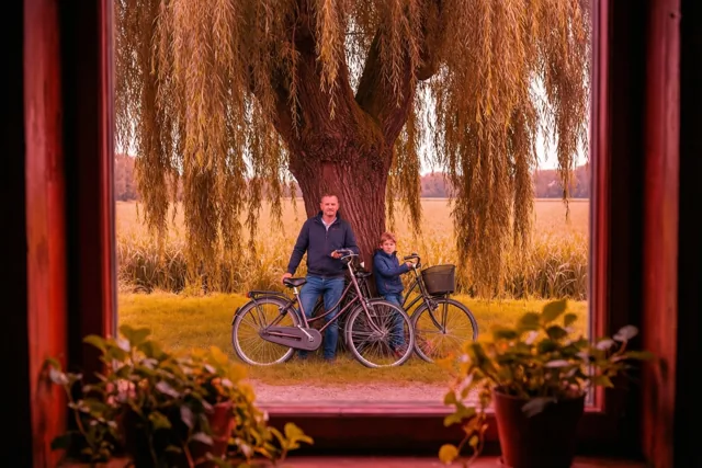 Door een rood raamkader met potplanten op de voorgrond zijn een man en kind te zien die naast hun fietsen tegen een knotwilg leunen in een polderlandschap met warme gouden en rode tinten.