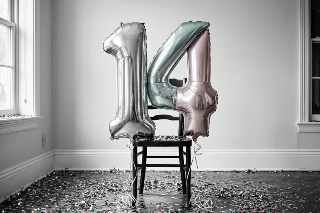 Twee grote folieballonnen vormen 14 aan een donkere houten stoel in een lichte, sobere kamer met confetti en ramen aan beide kanten.