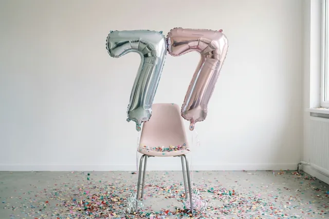 Twee grote folieballonnen in de vorm van 77 hangen aan een roze stoel in een lichte kamer met een witte muur, een raam en confetti op de vloer.