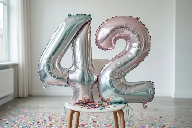Zilverkleurige en pastelroze folieballonnen in de vorm van 4 en 2 zijn vastgebonden aan een witte stoel, met kleurrijke confetti op de vloer.
