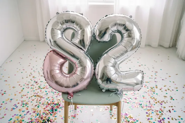 Twee grote folieballonnen in de vorm van een 8 en een 2 hangen aan een stoel in een lichte kamer met kleurrijke confetti en zilveren, pastelroze en witte tinten.