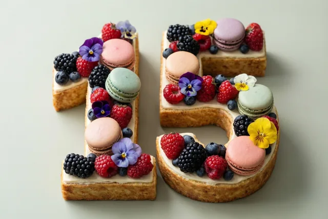 Een taart in de vorm van 15 is versierd met frambozen, bramen, blauwe bessen, macarons en eetbare bloemen op een effen lichtgroene ondergrond.