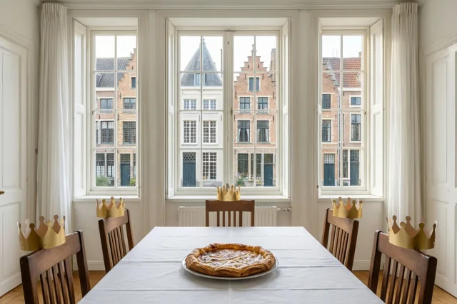 In een lichte eetkamer met witte muren en drie grote ramen staat een tafel met een galette en houten stoelen met papieren kronen, met uitzicht op Vlaamse bakstenen gebouwen.